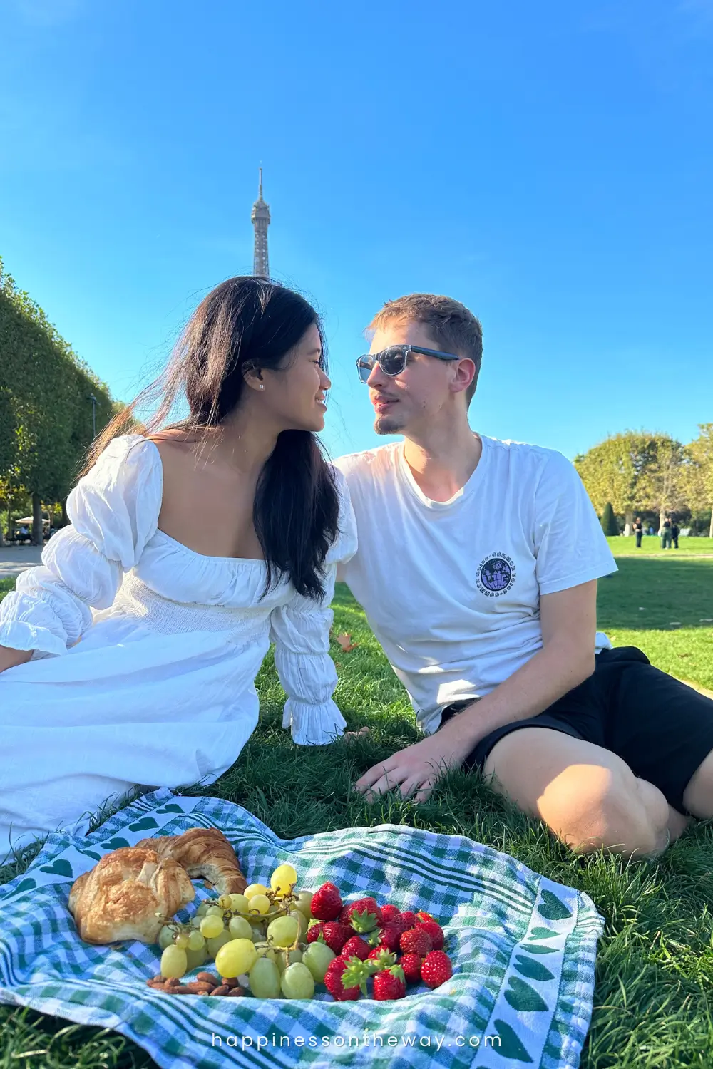 Me and Mat sharing a tender moment at a picnic at the Eiffel Tower, gazing into each other's eyes, with the woman dressed in a white puff-sleeved dress and the man in a casual white t-shirt and sunglasses. They're seated on the grass with a picnic setup featuring a croissant, grapes, strawberries, and almonds on a checkered cloth.