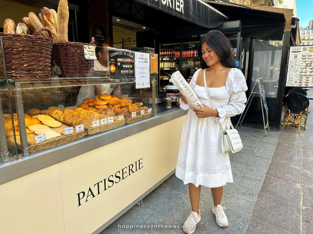 Me in a flowing white dress smiles as she reads a menu outside a traditional French patisserie. The bakery display is filled with an assortment of fresh bread and pastries, including golden croissants and baguettes, invitingly showcased behind the glass counter, with the patisserie sign in bold letters anchoring the scene.