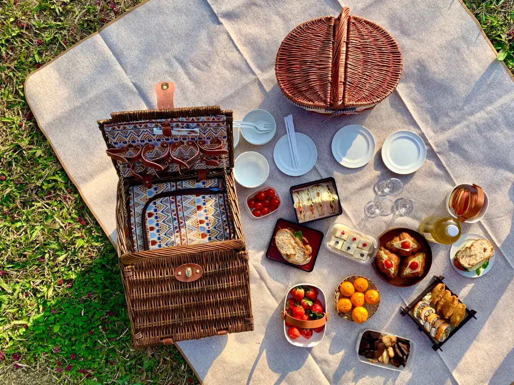 An overhead shot of an elegantly arranged picnic tour Eiffel on a beige blanket, featuring an ornate wicker basket, a selection of dishes with fresh strawberries, cherry tomatoes, oranges, and assorted sandwiches, complemented by glasses and a bottle of wine, all basking in the warm glow of the sunlight, ready for a luxurious outdoor feast.