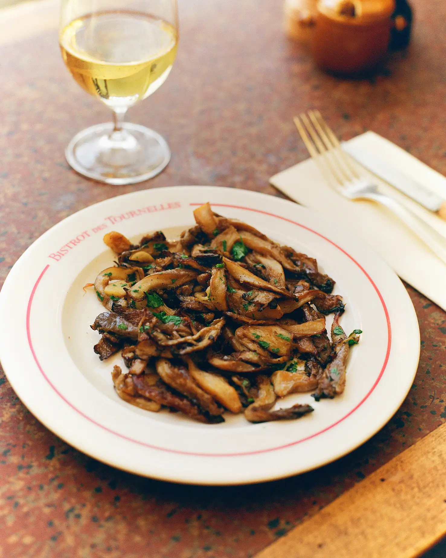 A plate of sautéed mushrooms served at Bistrot Des Tournelles in Paris, with a glass of white wine on a terrazzo table.