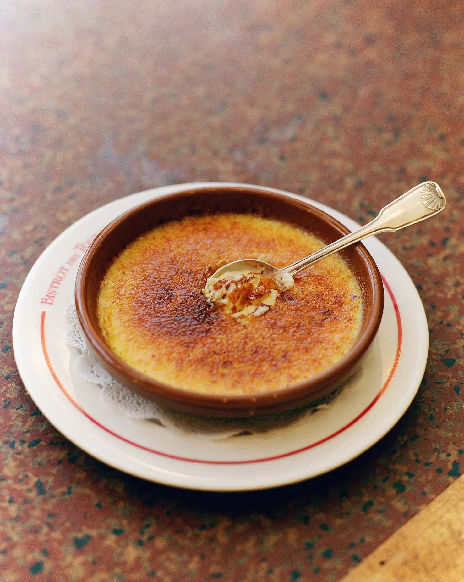 A classic crème brûlée with a caramelized top and a spoonful taken out, served at Bistrot Des Tournelles in Paris on a bistro-branded dish.
