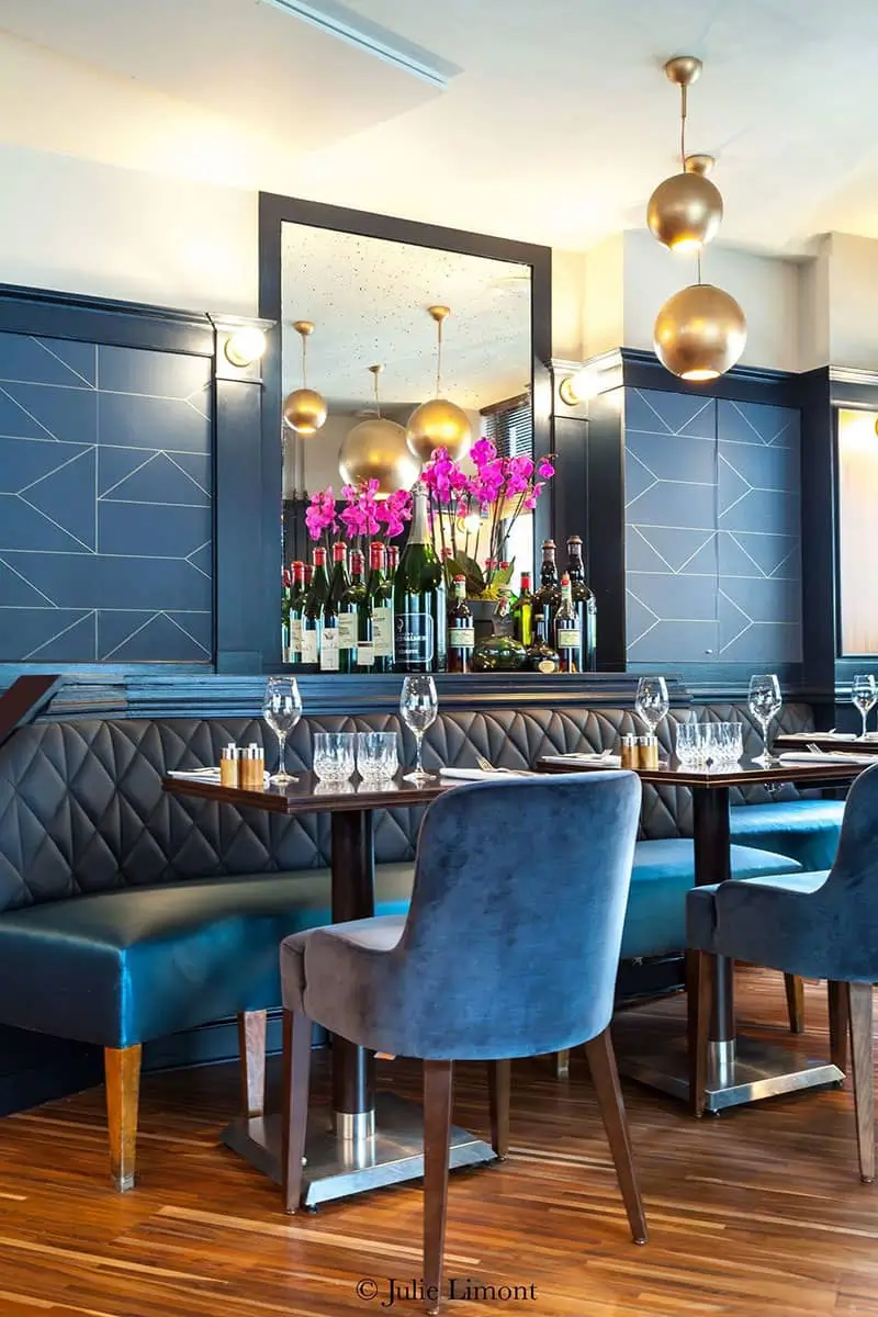 Chic dining area at Bistrot Marloe in Paris featuring blue velvet chairs, wooden tables, and modern gold pendant lights, with a large mirror reflecting vibrant pink orchids and wine bottles.