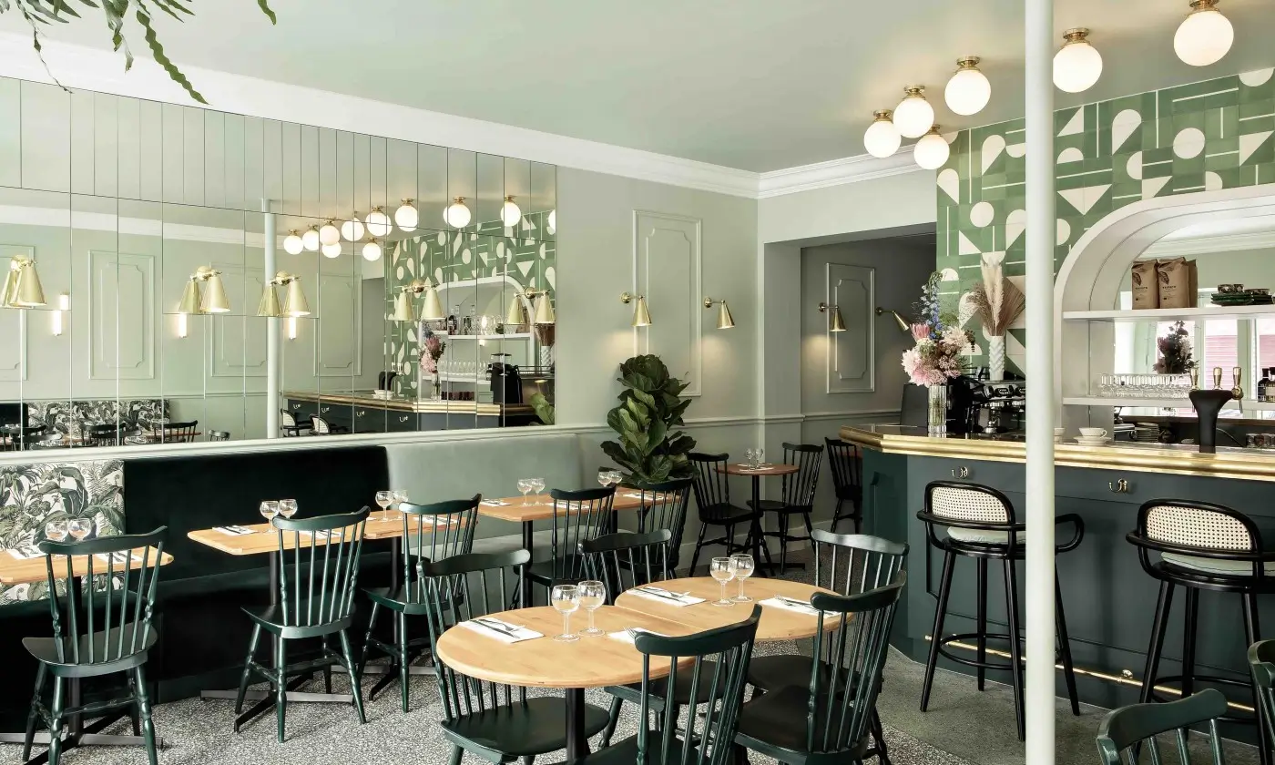 The interior of Bistrot Rougemont in Paris, featuring a serene color palette, green seating, patterned cushions, brass lamps, and a mirrored wall reflecting the inviting bar area.