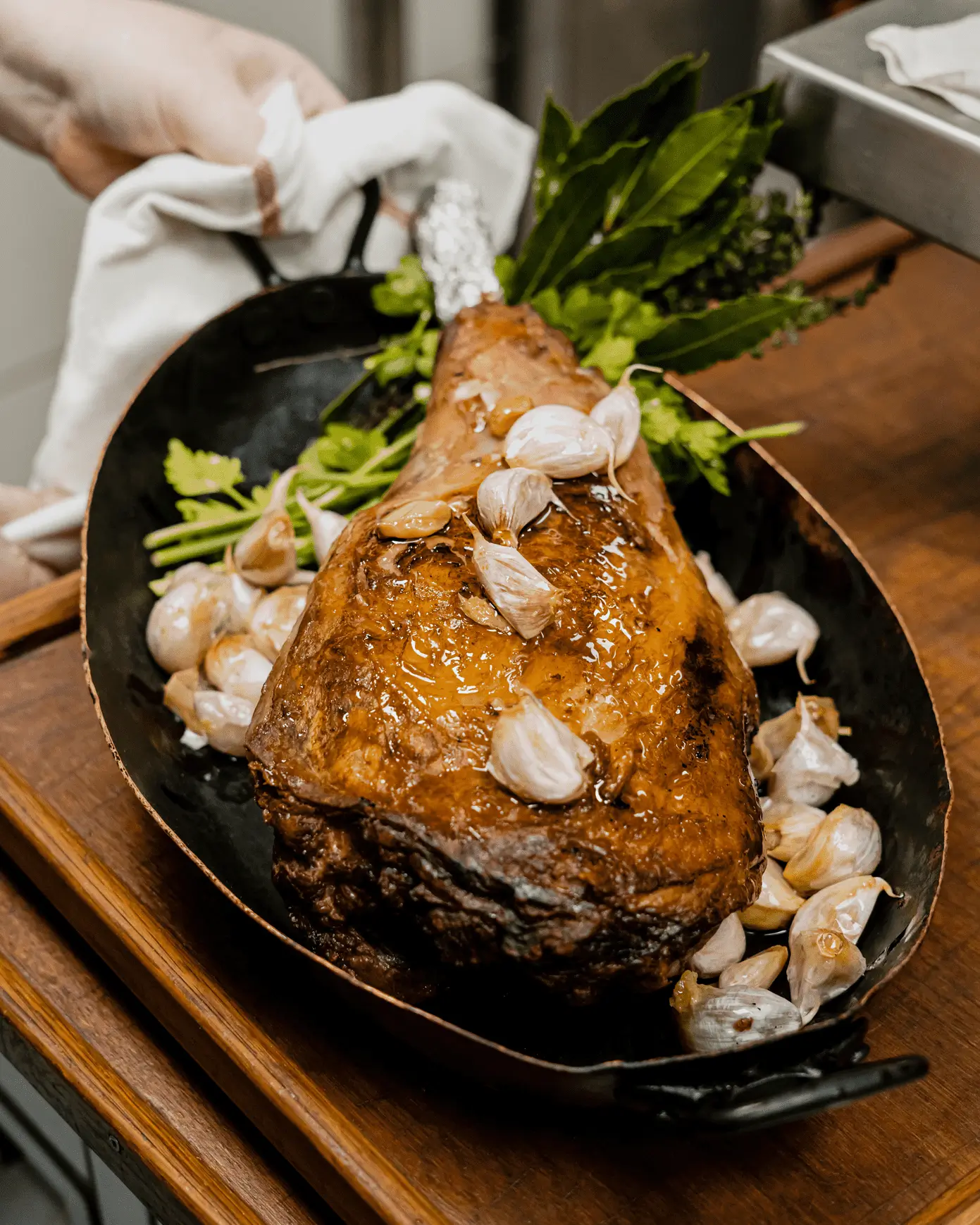 A succulent roasted leg of lamb garnished with whole garlic cloves, presented in a rustic skillet on a wooden surface, showcasing the classic cuisine at Chez Georges, a traditional French bistro in Paris.
