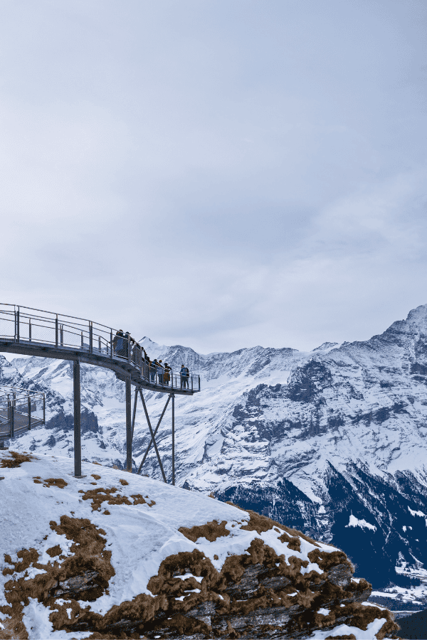 Visitors on the Cliff Walk at Grindelwald First, a metal walkway perched above a dramatic alpine landscape with snow-dusted mountains. One of the reasons why Grindelwald First is worth visiting.