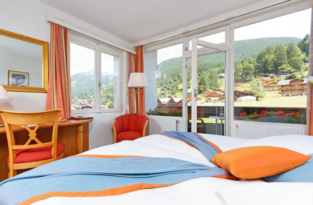 A cozy room of Derby Swiss Quality Hotel with a comfortable bed, warm lighting, and large windows offering a picturesque view of Grindelwald village.