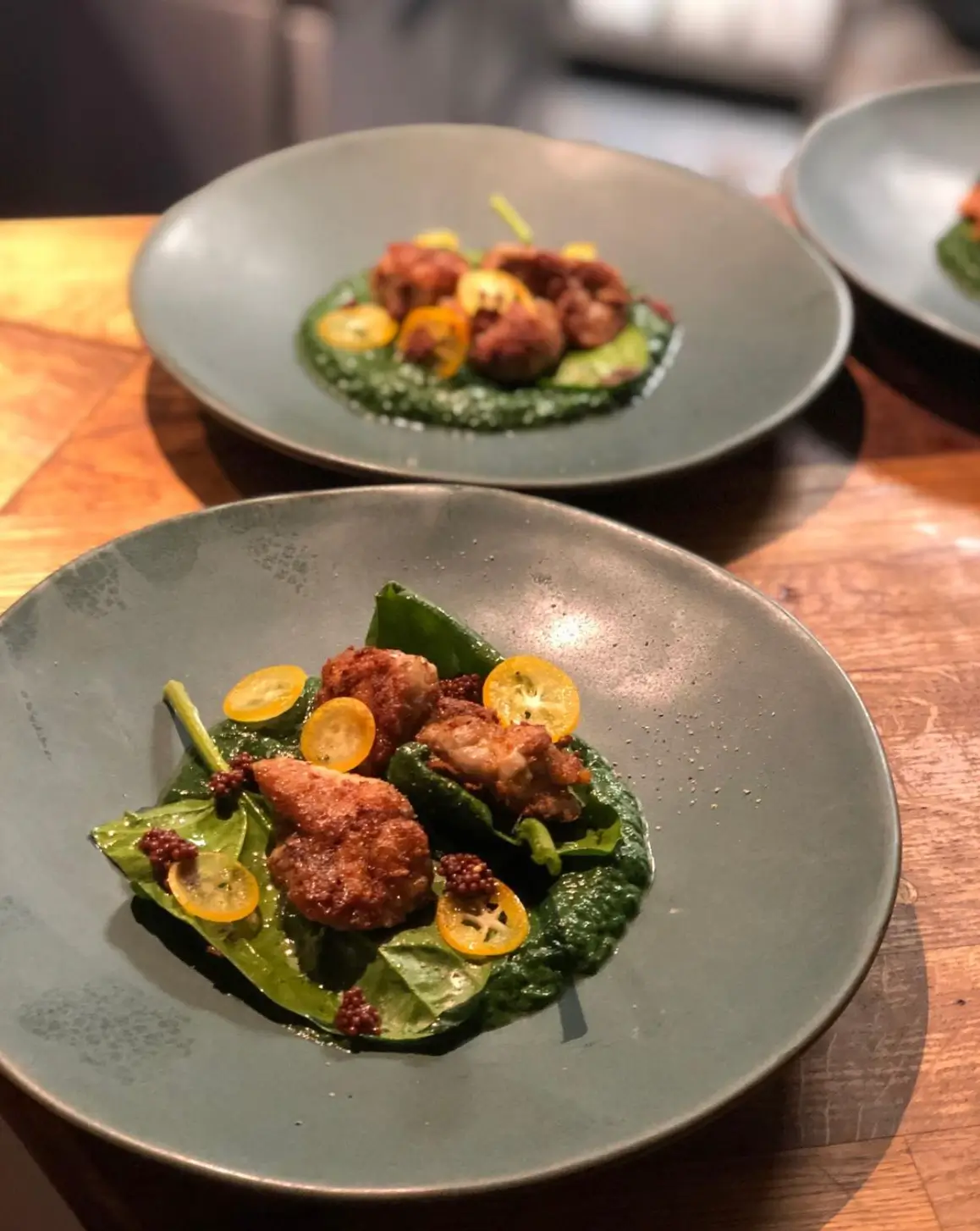 A gourmet dish featuring fried morsels on a bed of vibrant green sauce and garnished with kumquat slices, presented on a slate-colored plate at Des Terres, Paris.