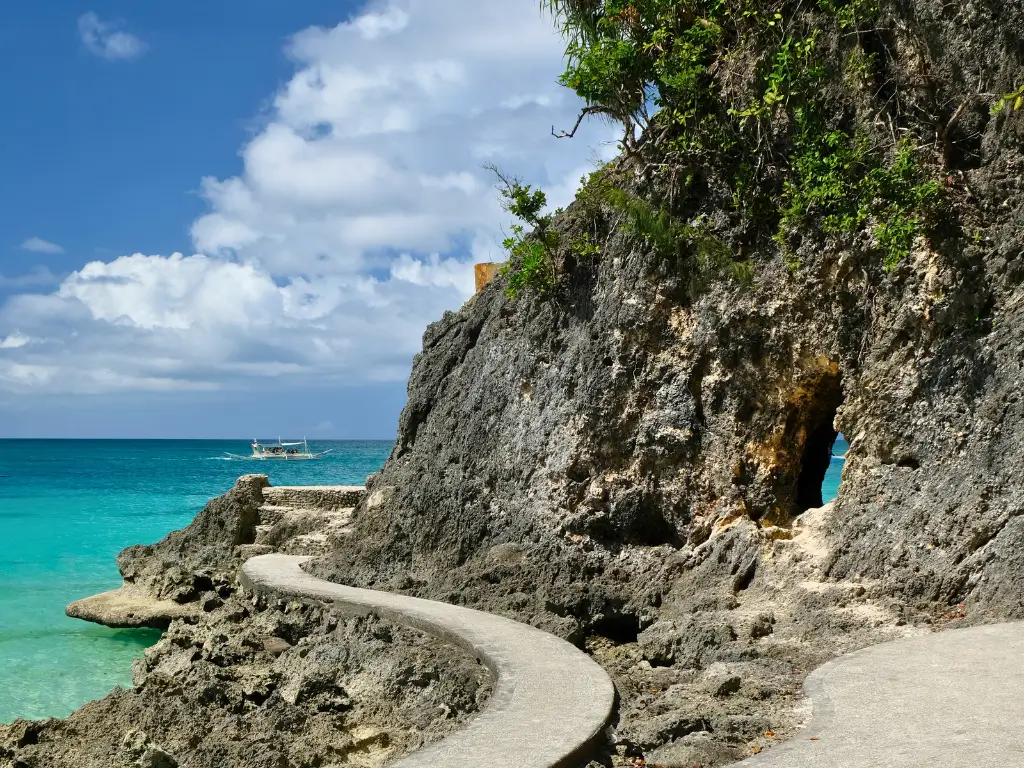 Winding footpath along the rocky cliffside of Diniwid Beach, leading to turquoise waters with a traditional boat in the distance, a scenic part of Boracay land tour destinations.