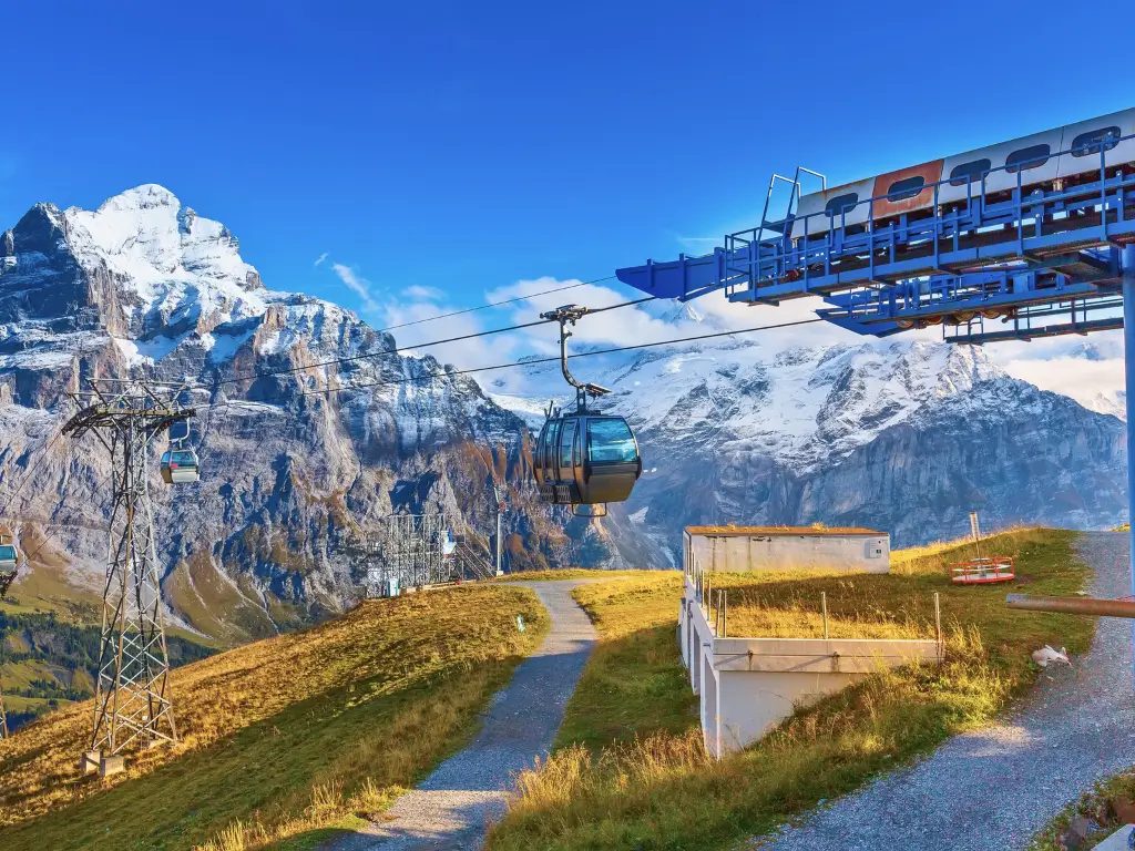A cable car gliding smoothly towards the snow-capped peaks of Grindelwald First under a brilliant blue sky, showcasing the adventure and stunning alpine scenery that make Grindelwald a worthwhile visit.