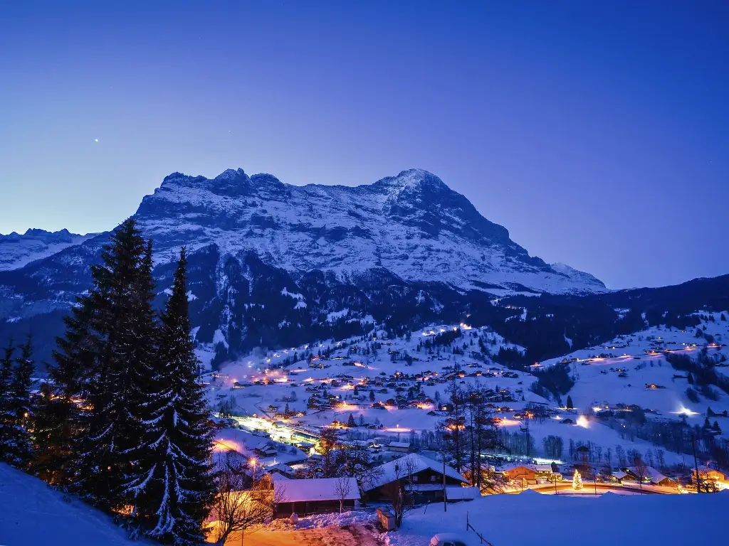 Nighttime in Grindelwald with twinkling lights from the village below illuminating the snowy landscape, all set against the dramatic silhouette of the Eiger mountain.