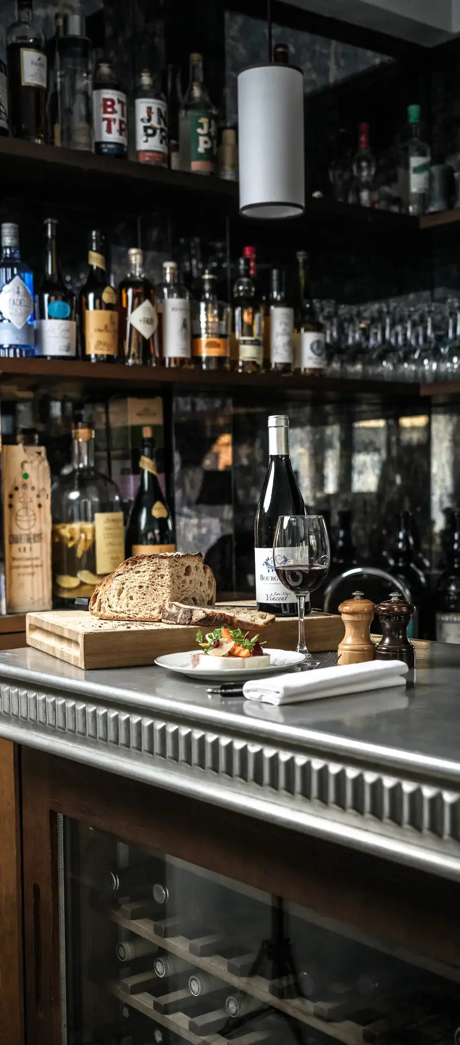 An intimate bar setting at Janine Bistro in Paris with a selection of liquors on shelves, freshly baked bread on a cutting board, and a glass of red wine in focus on the counter.