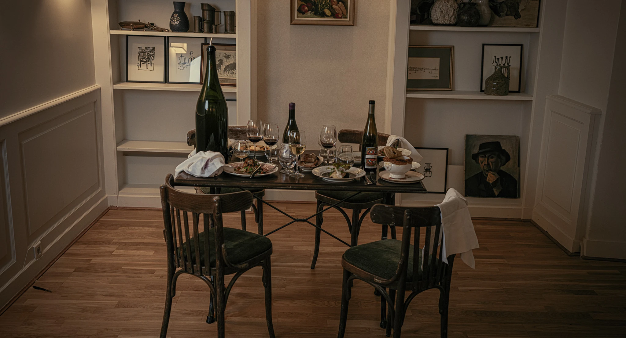 A rustic dining setup at Janine Restaurant, a French bistro in Paris, with a meal for two, multiple wine glasses, and artfully arranged wall shelves with various decorative items.