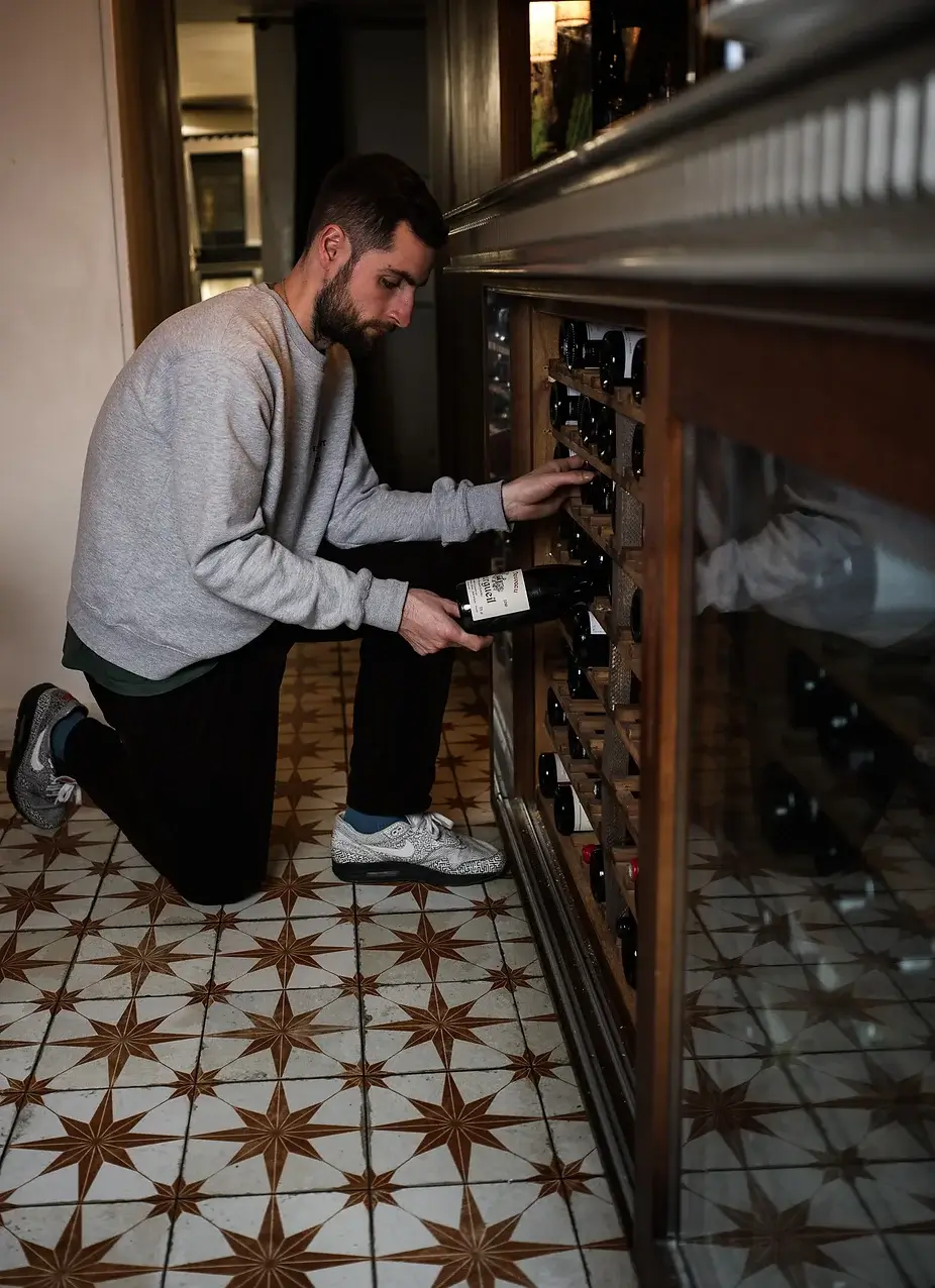 A man carefully selecting a bottle of wine from a wine fridge at Janine Restaurant in Paris, with traditional patterned floor tiles.