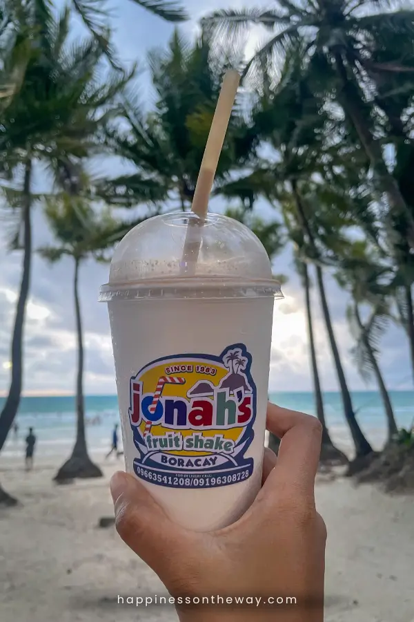 Hand holding a Jonah's fruit shake with a rice straw, a famous refreshment in Boracay, with palm trees and the beach in the background.