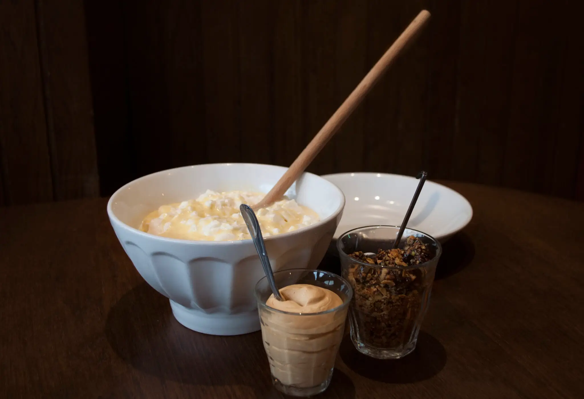 Famous creamy rice pudding from L'Ami Jean in Paris, served in a large white bowl with a wooden spoon, accompanied by a glass of whipped cream and a jar of granola.