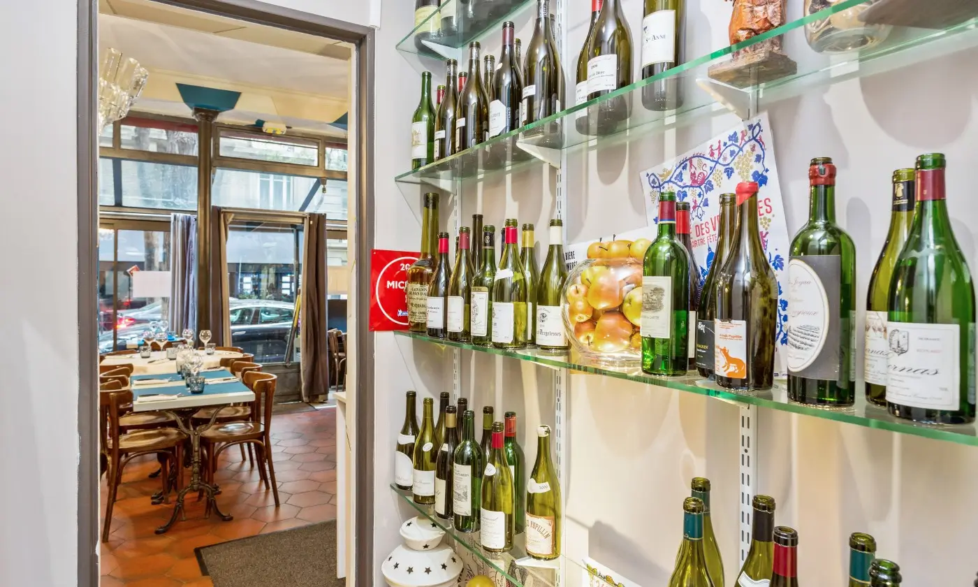 Inside Le Bistrot du Maquis, Paris, with wine bottles on shelves beside a dining area with set tables and chairs.