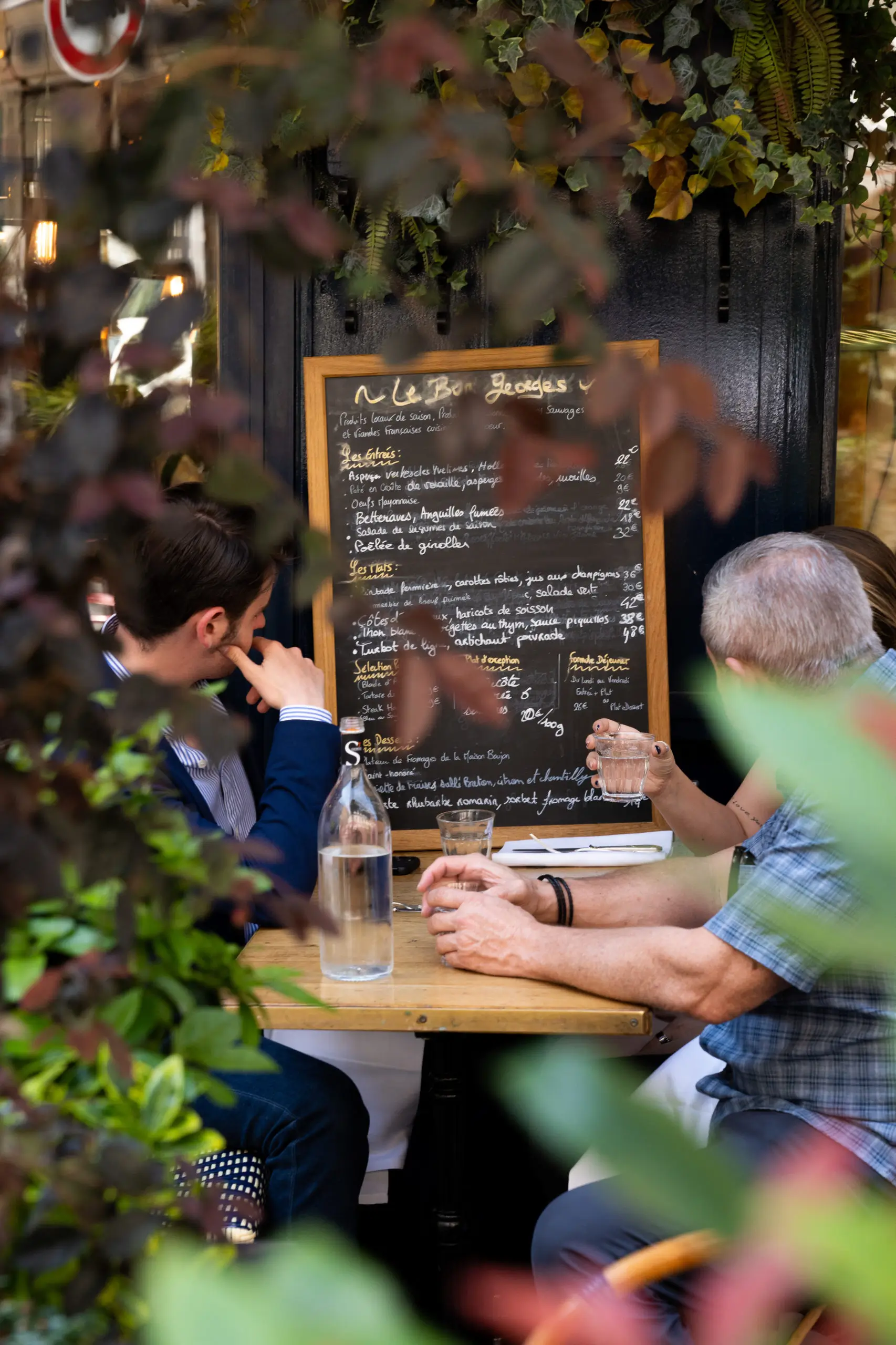 Two customers seated at an outdoor table of Le Bon Georges in Paris, reviewing a chalkboard menu framed by lush greenery.