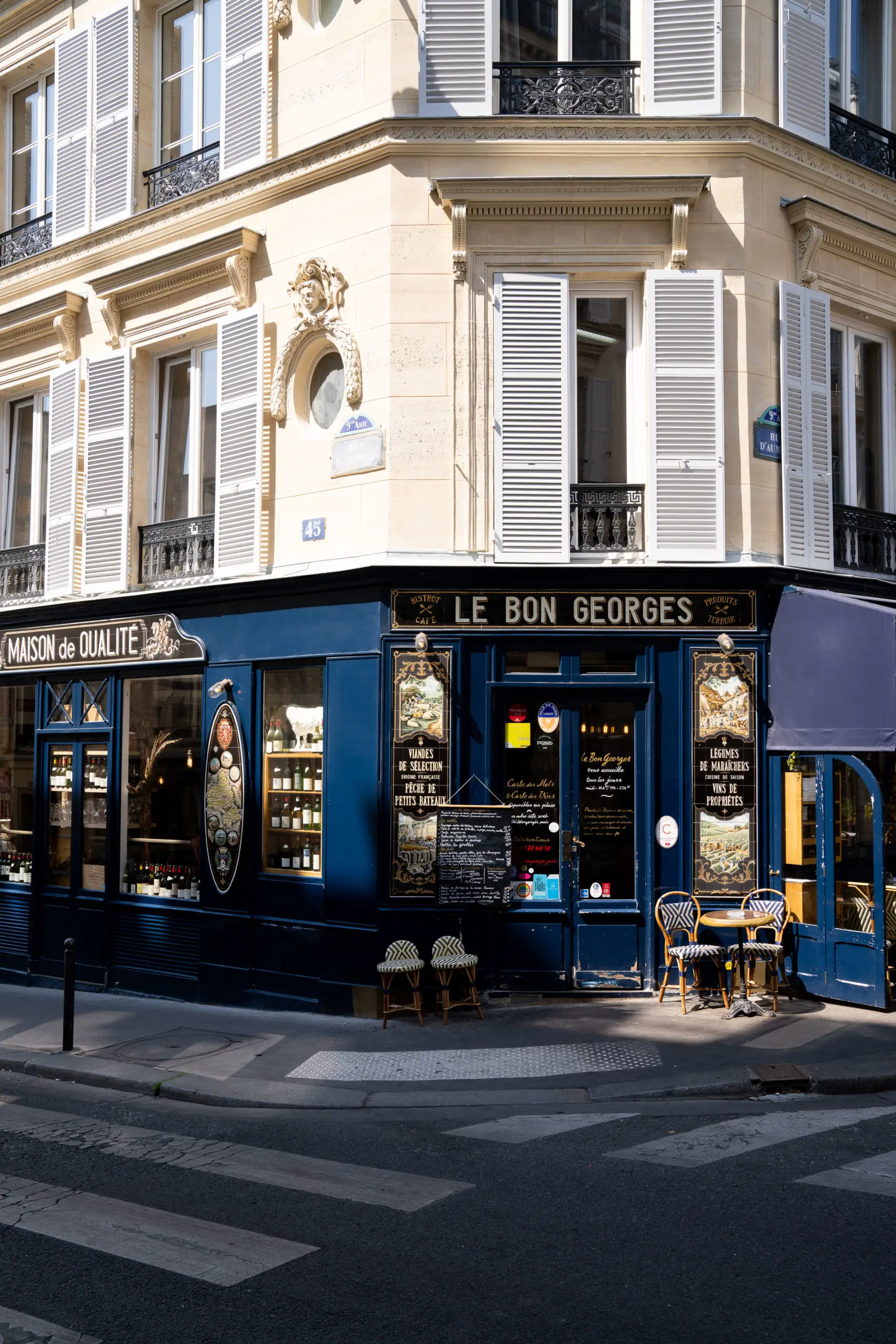 The corner of Le Bon Georges, a classic bistro in Paris, with a navy blue exterior, golden lettering, bistro chairs outside, and a traditional Parisian building facade.