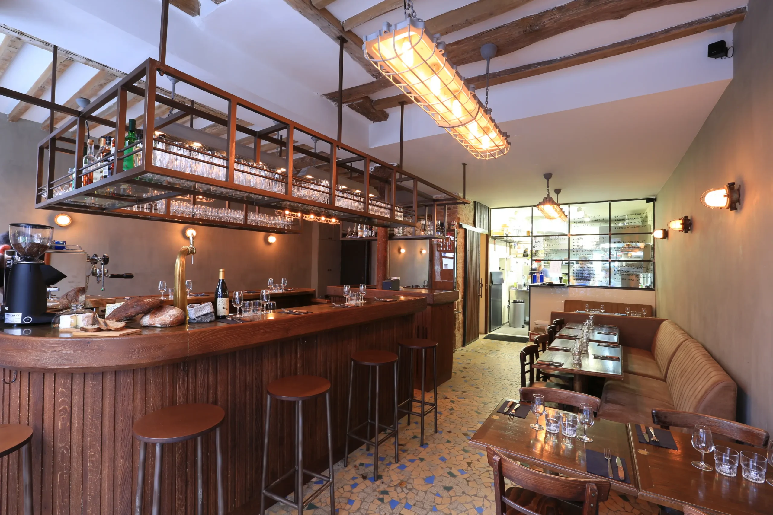 The rustic and inviting interior of Le Grand Bain Bistro in Paris, with an exposed beam ceiling, tiled flooring, a cozy bar area with hanging glasses, and wooden tables ready for guests.