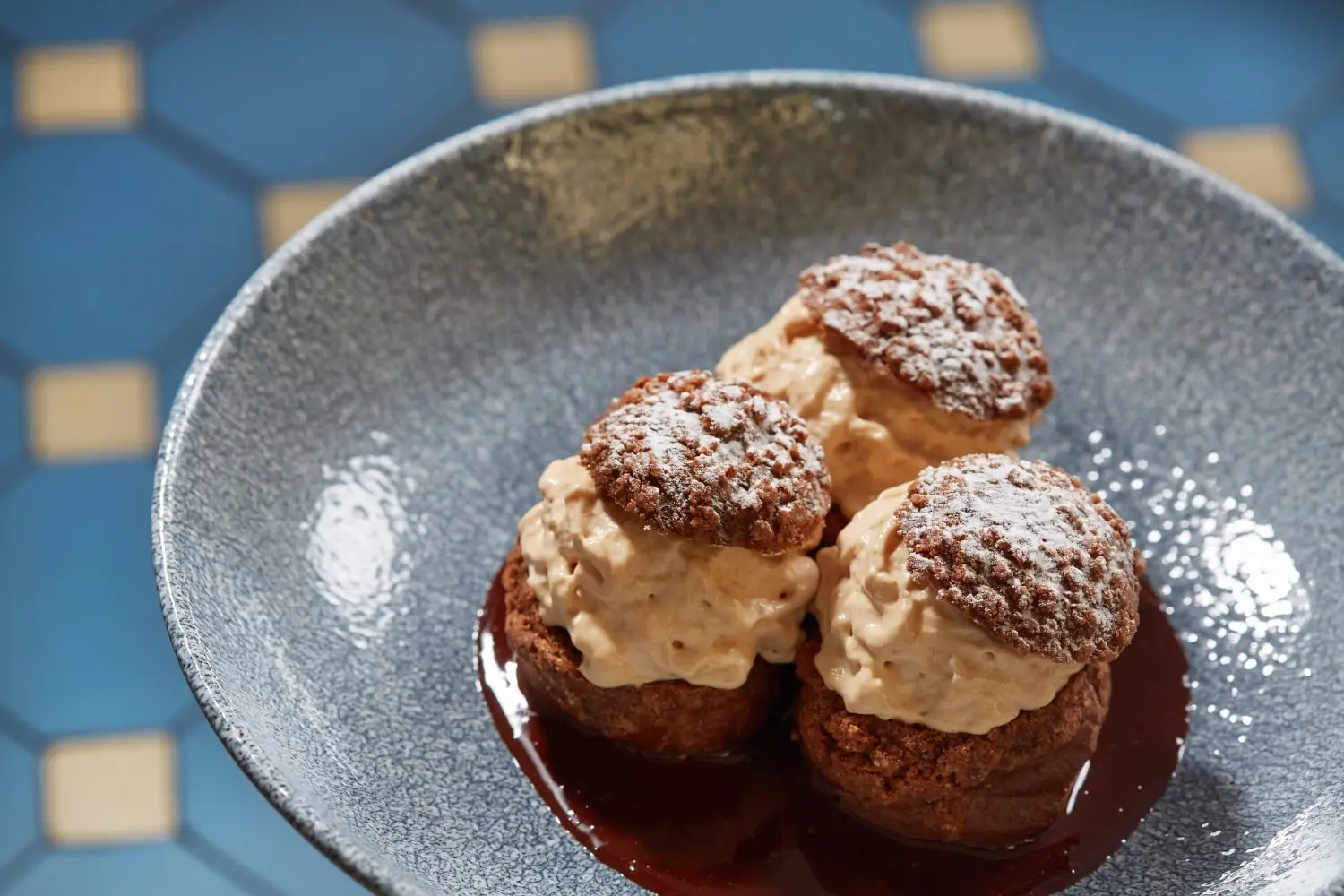 Gourmet profiteroles filled with cream and drizzled with chocolate sauce, served on a dark ceramic plate at Les Canailles Paris, ready to delight any dessert aficionado.