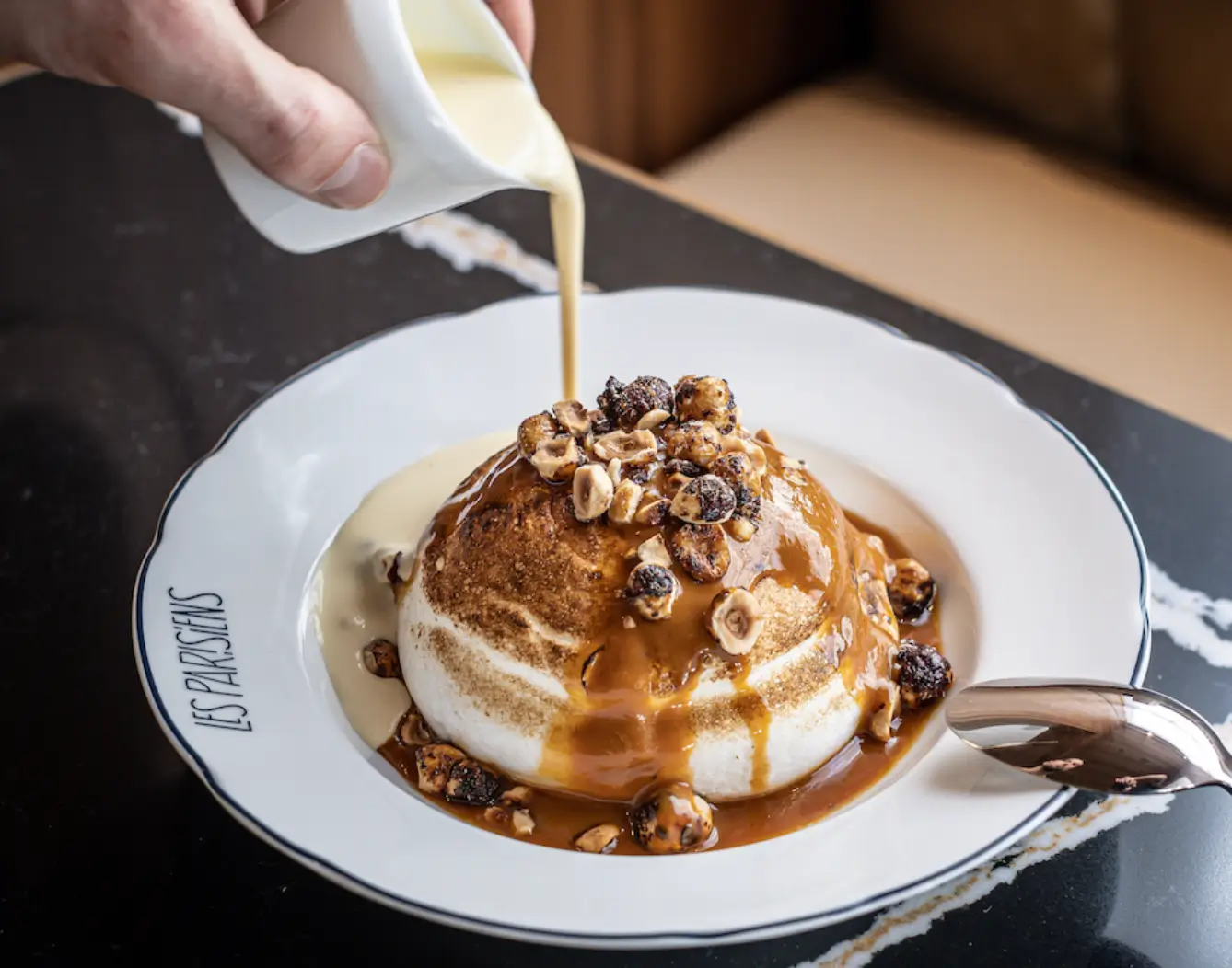 Dessert at Les Parisiens Paris, featuring a panna cotta topped with hazelnuts and caramel sauce, served on a branded plate.