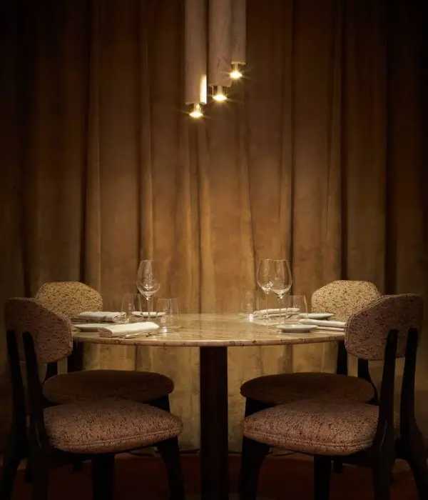 Intimate dining setting at Mallory Gabsi's restaurant in Paris, renowned for its affordable Michelin-star experience, featuring a cozy table set for two, elegant glassware, and a warm, golden glow from a modern chandelier, all against a soft, draped curtain background.