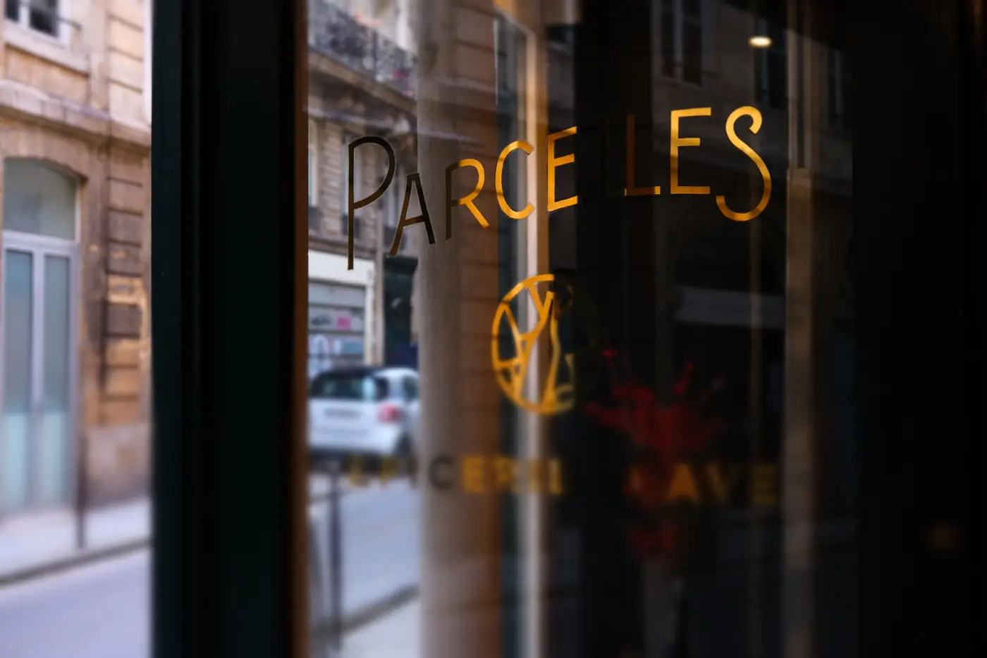 View through the glass window of Parcelles in Paris with the restaurant's name in gold lettering, reflecting the bustling street outside.
