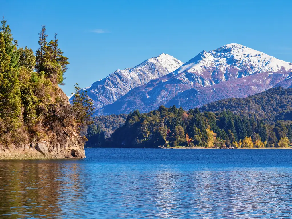 Tranquil blue waters meet a lush shoreline with snow-capped peaks in the background in Patagonia, an ultimate retreat for slow tourism destinations.