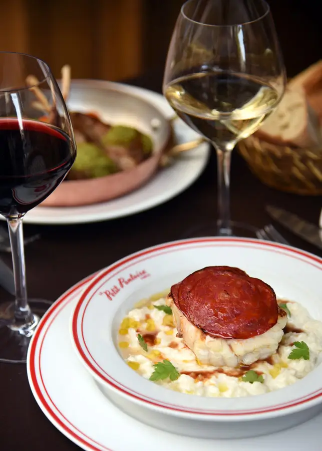 A gourmet plate of lotte with chorizo served at Le Petit Pontoise, paired with red and white wine, highlighting the bistro's fusion of French culinary traditions.