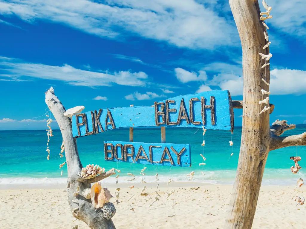 Wooden sign for 'Puka Beach Boracay' set against a clear blue sky on white sand, highlighting a popular stop among Boracay land tour destinations.