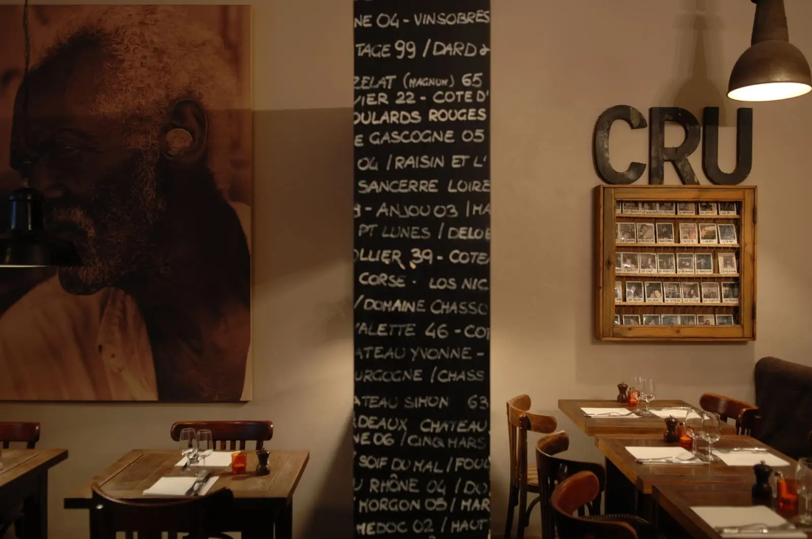 Warmly lit dining space at Cinq-Mars bistro in Paris, with tables ready for guests and a wall featuring a wine list, a large 'CRU' sign, and a framed collection of vintage photographs.