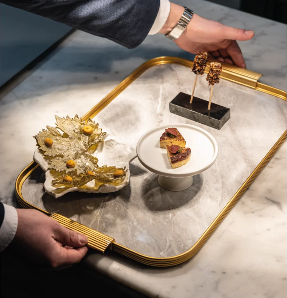 An elegant dish presentation at Restaurant Omar Dhiab, one of the affordable Michelin star restaurants in Paris, with a person's hand reaching for a skewer of decadent treats on a golden tray, beside a plate of seared meat garnished with herbs