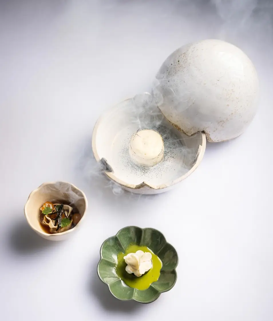 An immersive dining experience at Restaurant Pantagruel in Paris, where a culinary trio includes a smoky, spherical dome uncovering a creamy dessert, a side dish in an elegant petal-shaped bowl, and a bright green sauce with a dollop of foam, all presented on a serene white background.