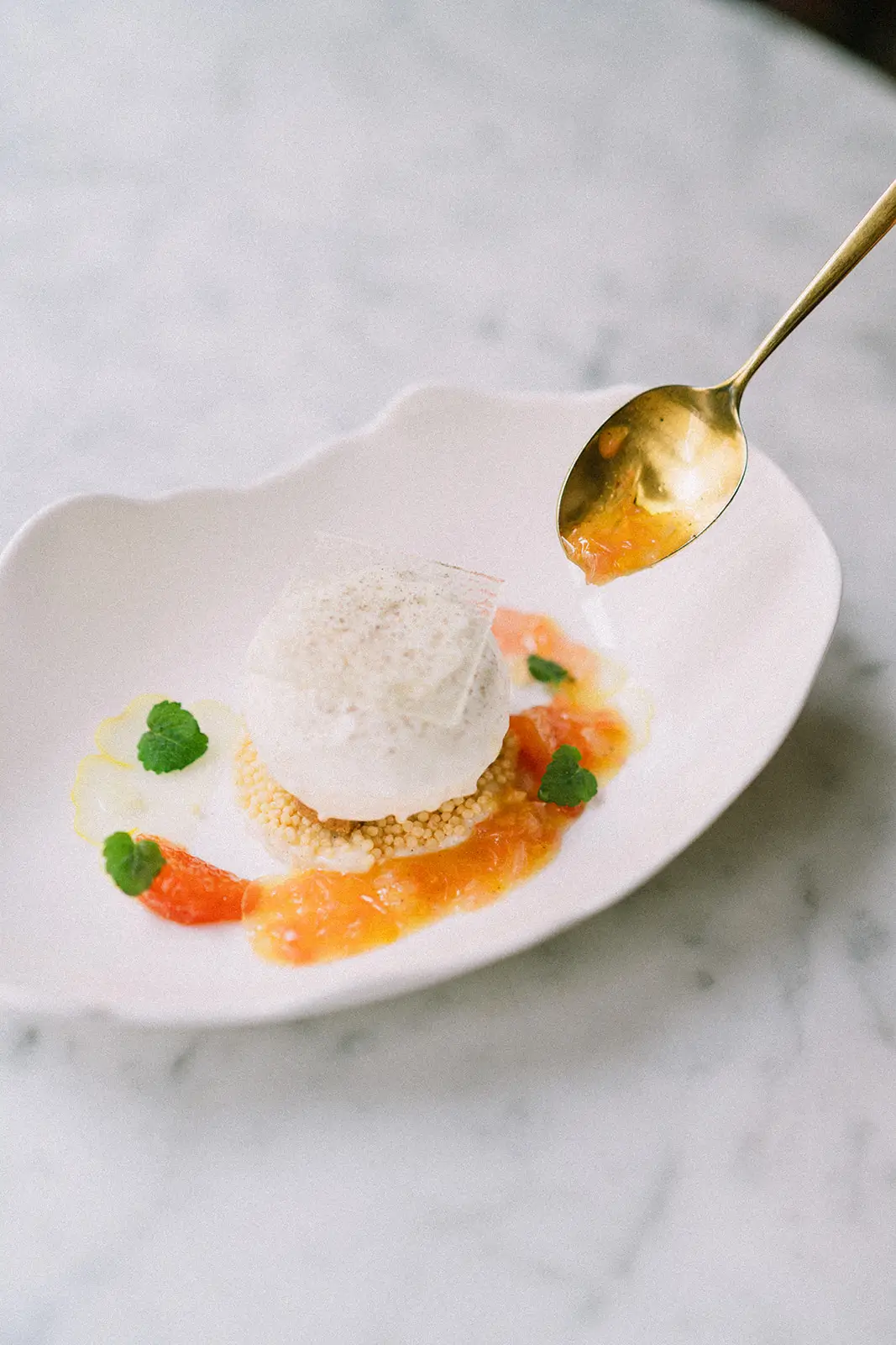 A delicate dessert at Virtus, a testament to the affordable luxury of Paris's Michelin-starred restaurants, featuring a scoop of ice cream atop a crunchy base, surrounded by citrus segments and fresh mint, served with a gold spoon on a marble surface.