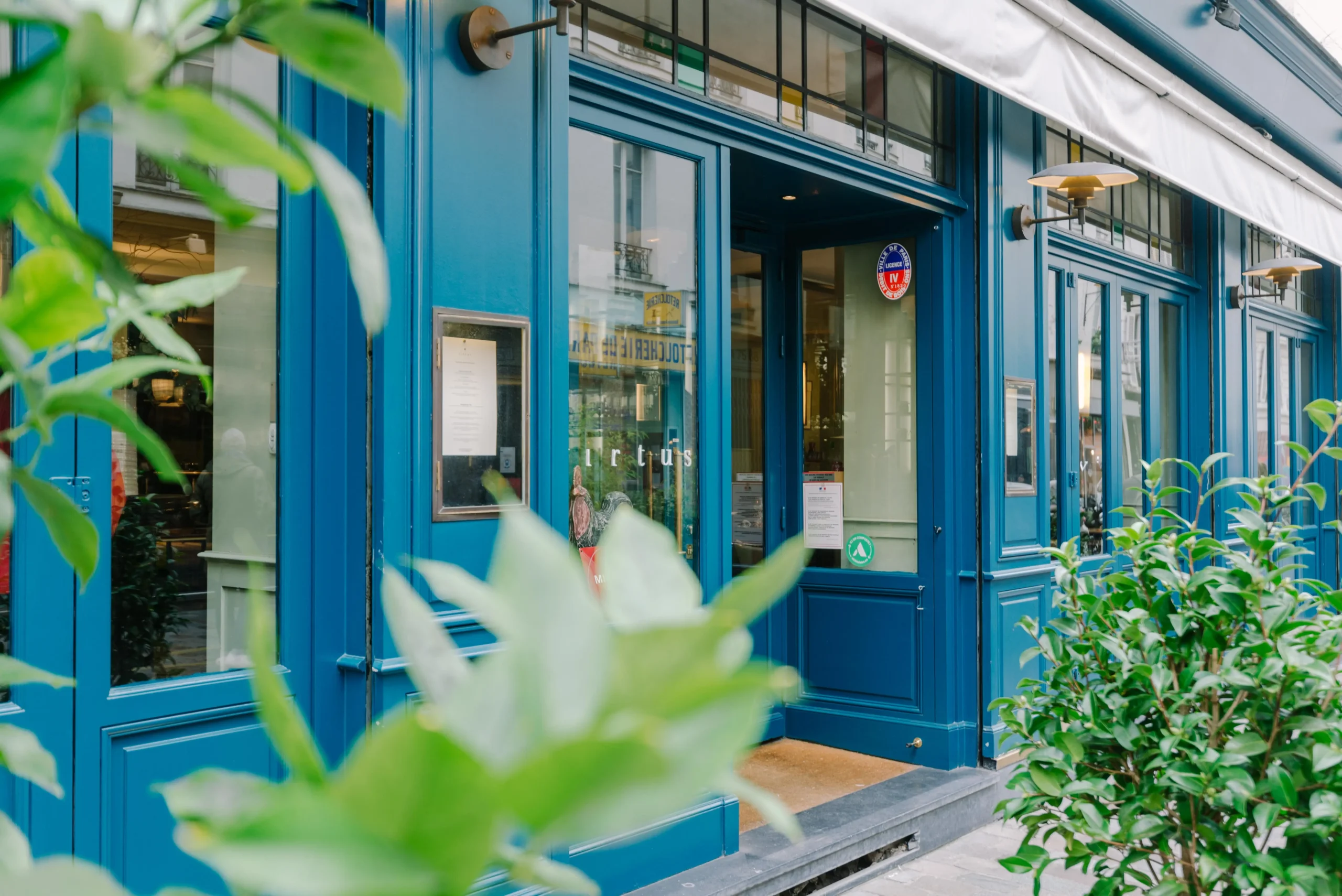 The charming blue facade of Virtus in Paris. It is recognized as best value Michelin restaurants Paris, invitingly opens to gourmet enthusiasts, framed by lush greenery in a vibrant street setting.