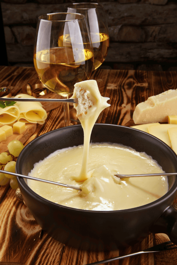 Traditional Swiss cheese fondue in a black pot, with bread being dipped and stringy cheese stretching, accompanied by glasses of white wine.