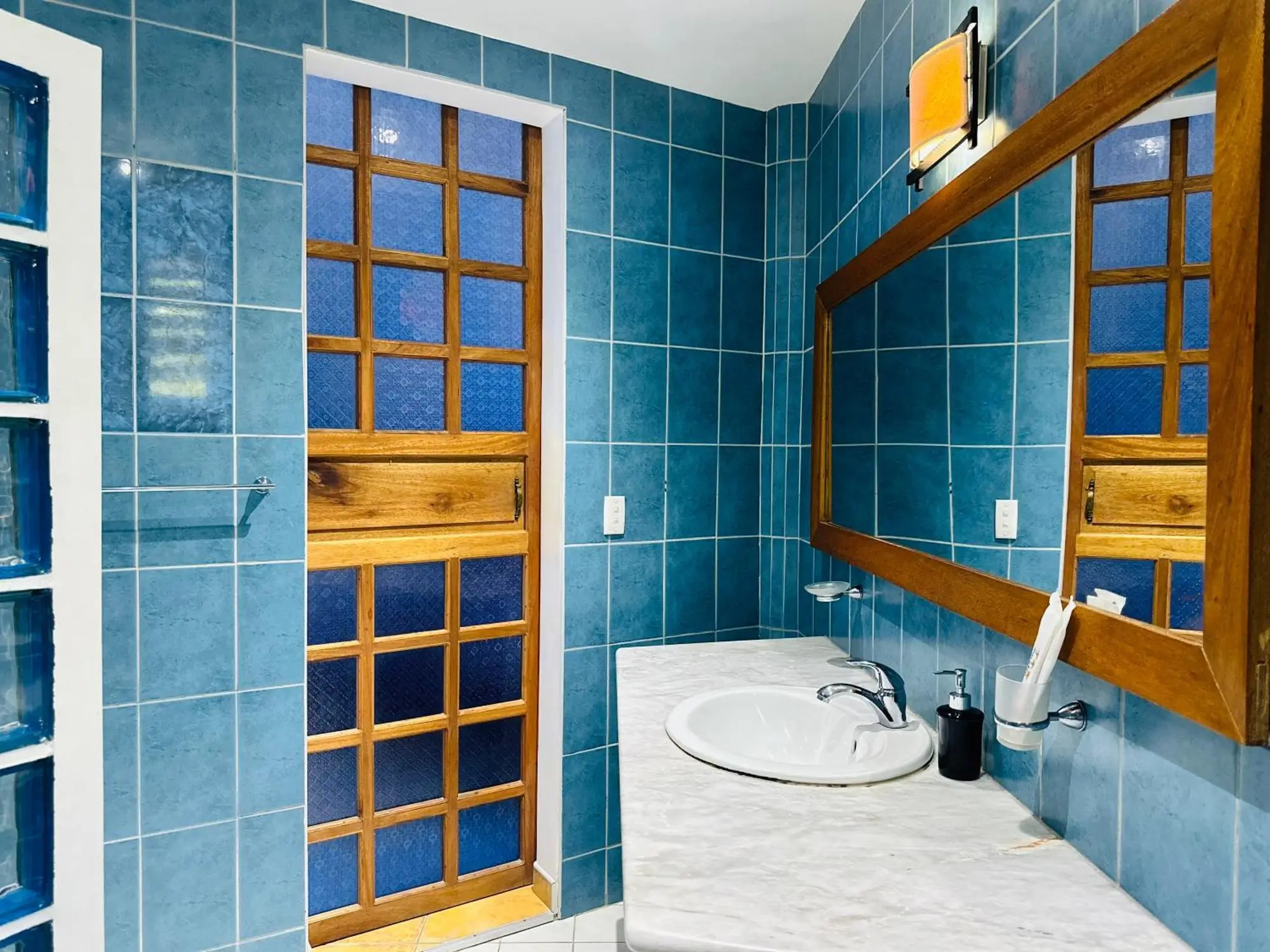 Bathroom in Aissatou Beach Resort featuring bright blue tile walls with a wooden door and frame, a white sink set on a marble countertop, and a large mirror reflecting a tastefully simple design.