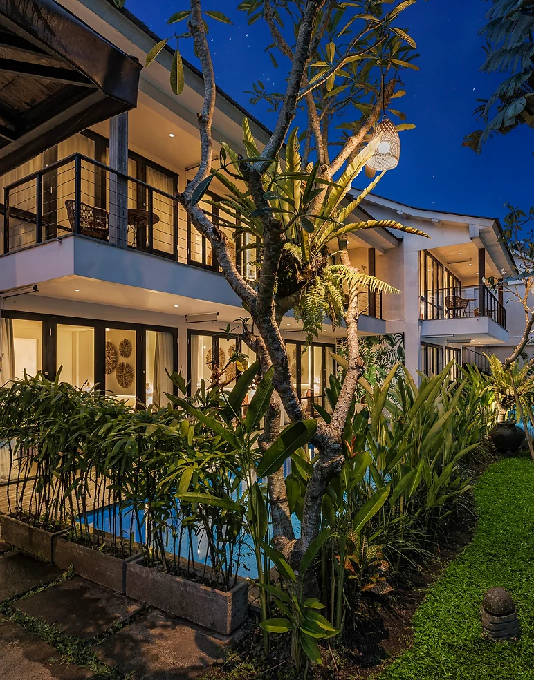 Night view of Ayurvedagram Bali, a modern wellness retreat featuring multi-story buildings with balconies, surrounded by vibrant tropical plants and an inviting blue pool. 