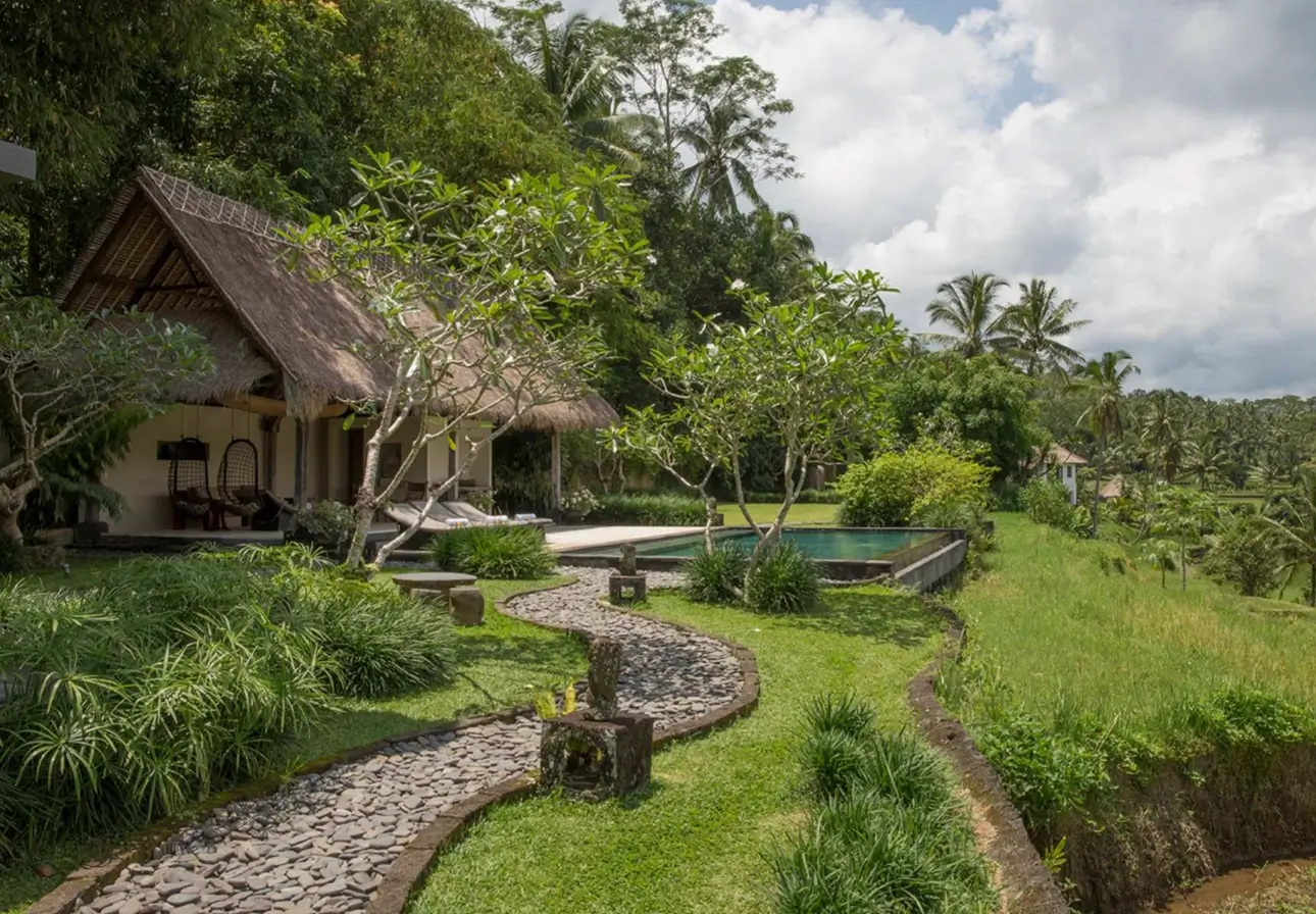A serene villa at Bliss Body Sacred Journeys wellness resort in Bali, featuring a thatched-roof bungalow with comfortable outdoor seating set amidst a lush garden. 
