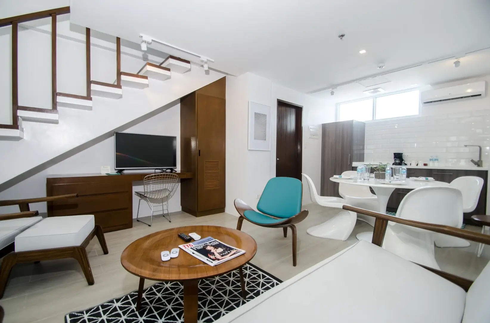 Modern loft-style room at Coast Boracay, featuring a minimalistic design with a TV, a small dining area, and an open stairway leading to the upper level. The room combines white walls and dark wood accents, creating a contemporary, inviting space.