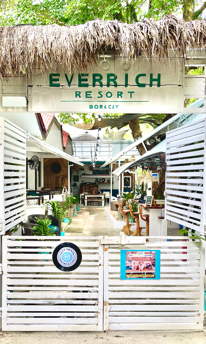 Entrance view of Everrich Boutique Resort in Boracay featuring a white gated entry with green signage under a thatched roof, leading into a vibrant, plant-decorated alley.