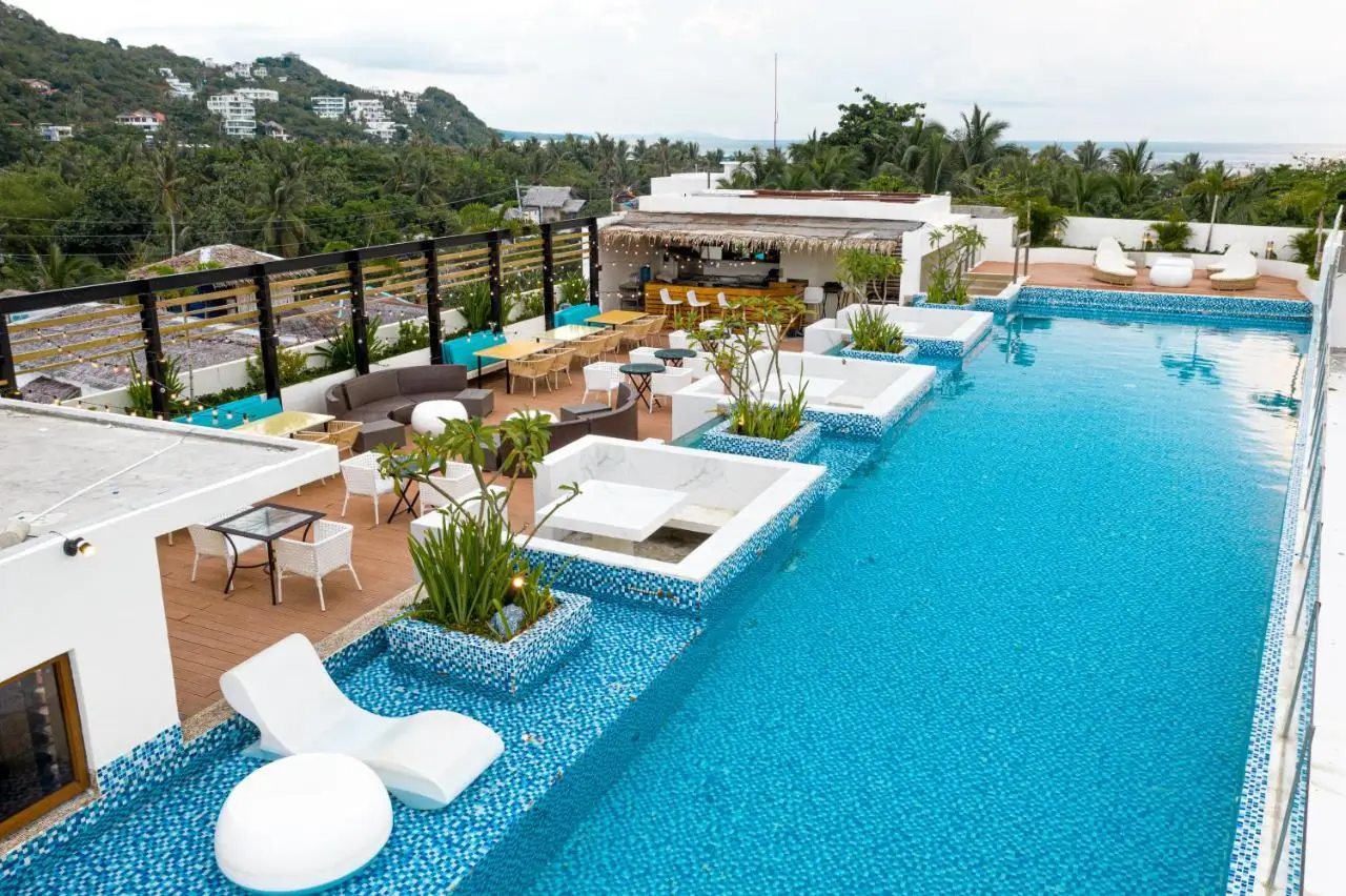 Luxurious rooftop swimming pool at Ferra Hotel in Boracay, a boutique hotel in Boracay, featuring white loungers and built-in seating areas scattered around a blue-tiled pool. The area also includes a bar and casual dining setup, all overlooking a lush, tropical landscape with views of the ocean in the distance.