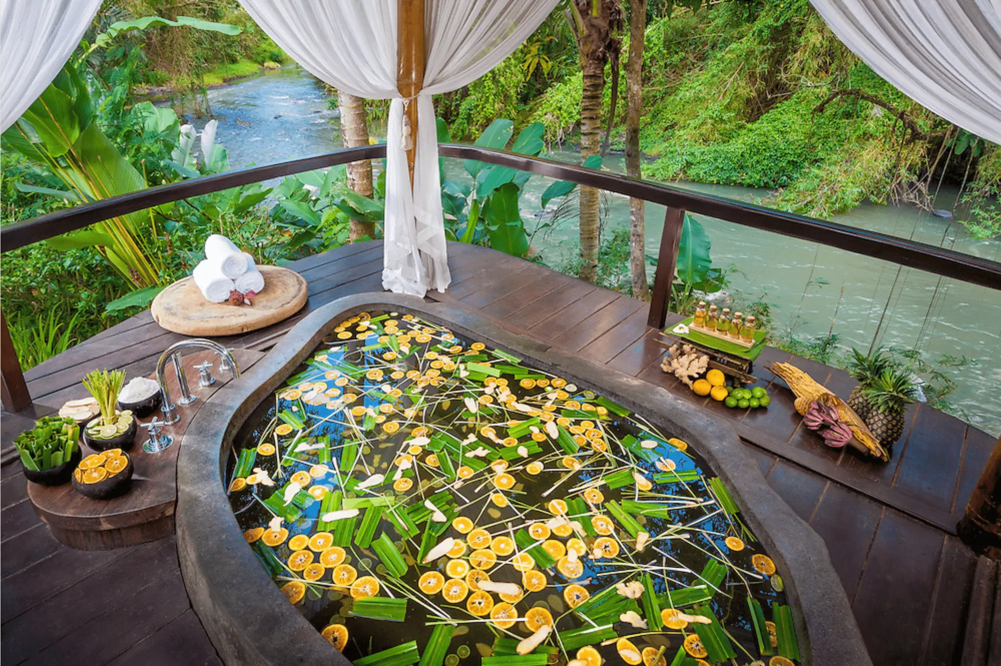 A detox spa bath at Fivelements Bali, filled with flowers and lemons floating on water, set on a wooden deck overlooking a lush river. 