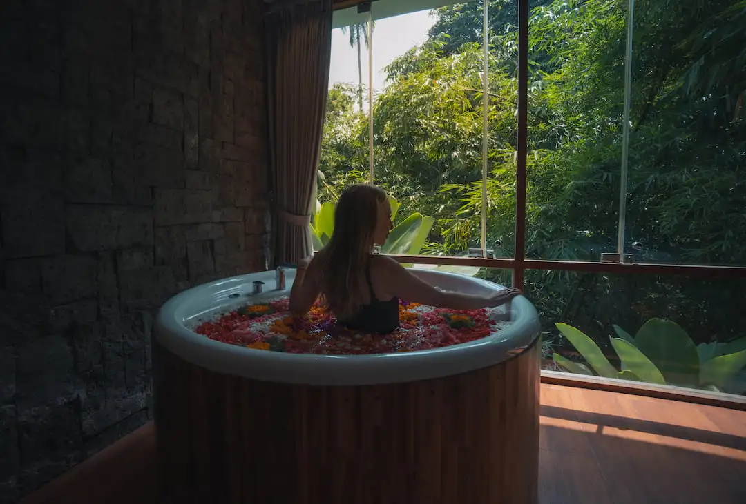 A woman relaxes alone in a flower bath in Bali, looking out a large window to a lush green view. The serene spa setting is complemented by natural light and stone walls.