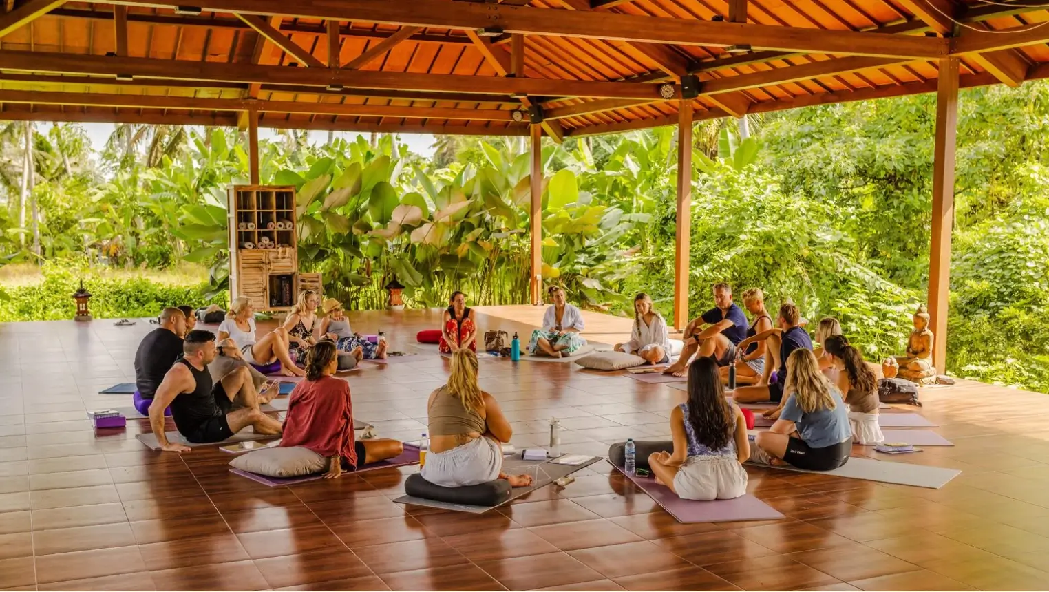 A diverse group of people seated on yoga mats in a spacious outdoor pavilion at Hot Stone Club Ubud Wellness Retreat, engaging in a wellness workshop.