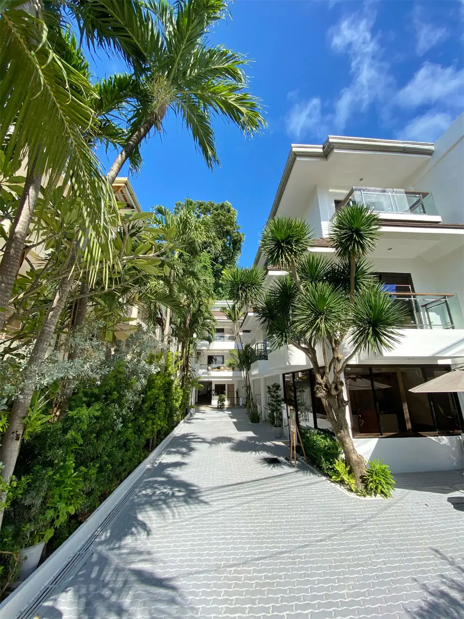 Paved walkway lined with lush green plants and palm trees inside Jony's Beach Resort in Boracay, leading to modern white buildings with balcony views.