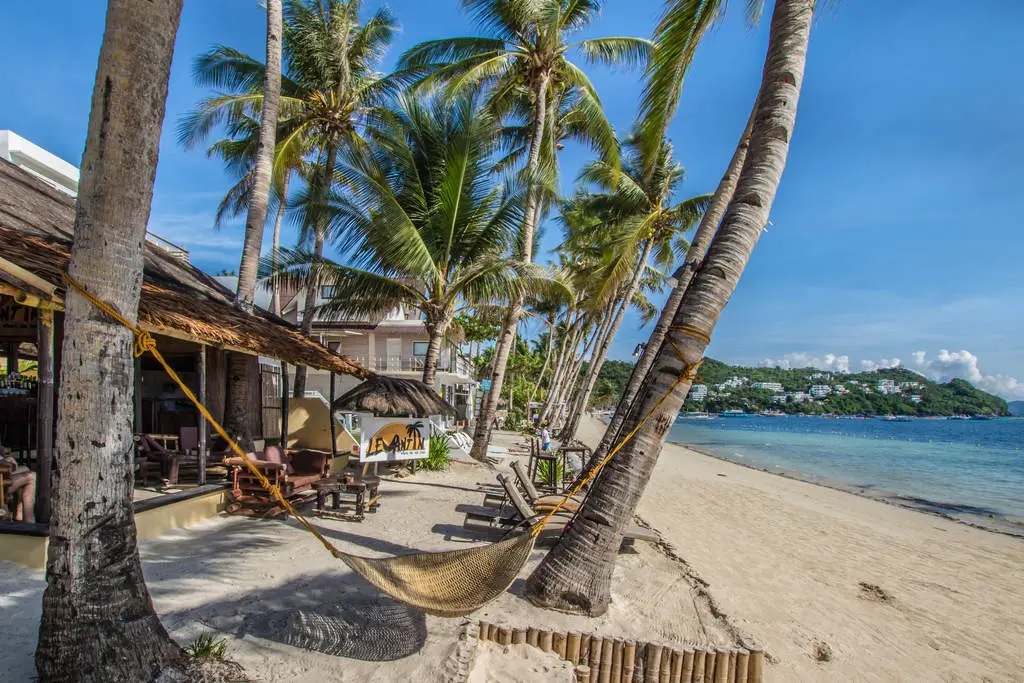 Beachfront at Levantin Boracay, a boutique resort in Boracay, featuring a sandy shoreline adorned with tall palm trees, a hammock tied between the trees, and rustic bars under thatched roofs, providing a serene sea view.