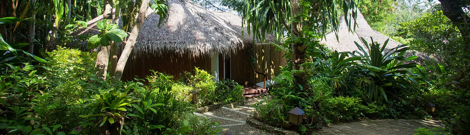 Secluded pathway leading to a private villa at Mandala Spa & Resort Villas, a boutique resort in Boracay. The villa is covered with a traditional thatched roof and surrounded by lush tropical greenery.