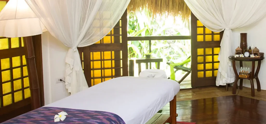Interior view of a spa treatment room at Mandala Spa & Resort Villas, featuring a massage table draped with a white cloth, surrounded by serene greenery visible through traditional Filipino style windows.