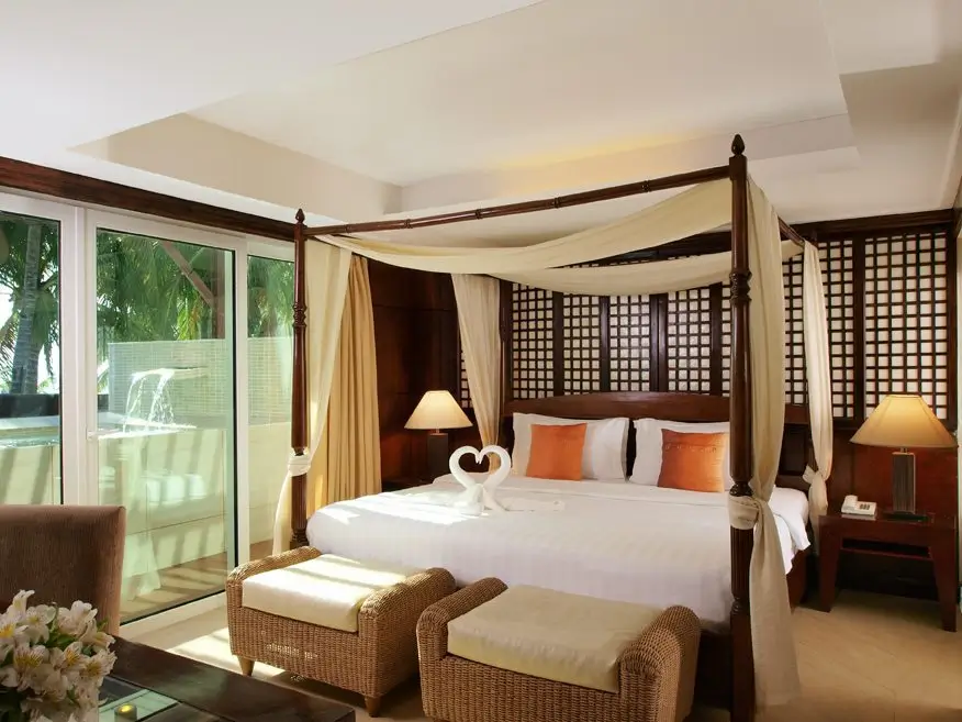 Elegant bedroom in Mandarin Bay Resort & Spa with a king-sized four-poster bed draped with light fabric, two cozy rattan chairs, and a large sliding glass door leading to a private balcony.